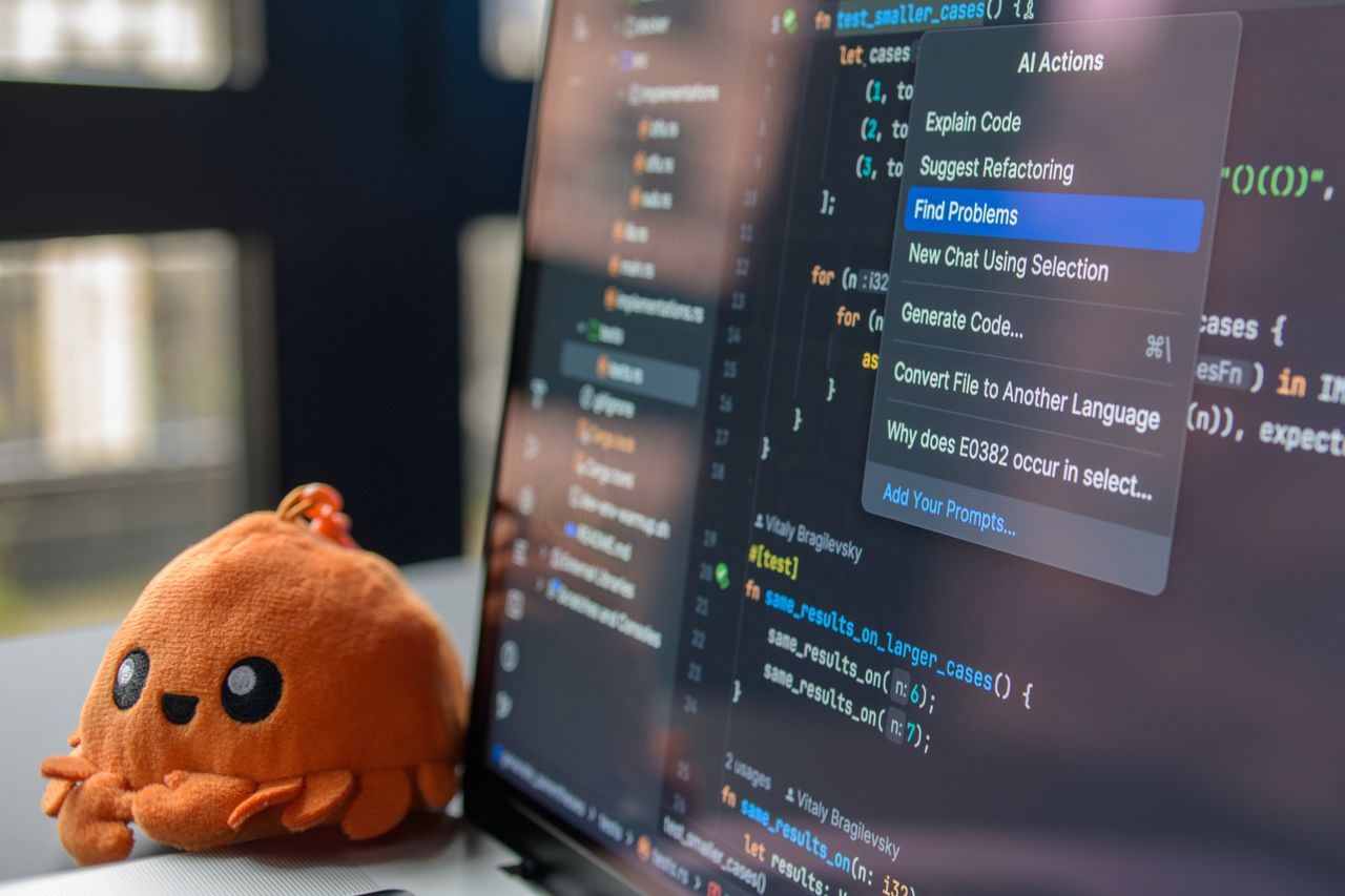 hero-img-01 A laptop screen showing a code editor with a cute orange crab plush toy beside it.